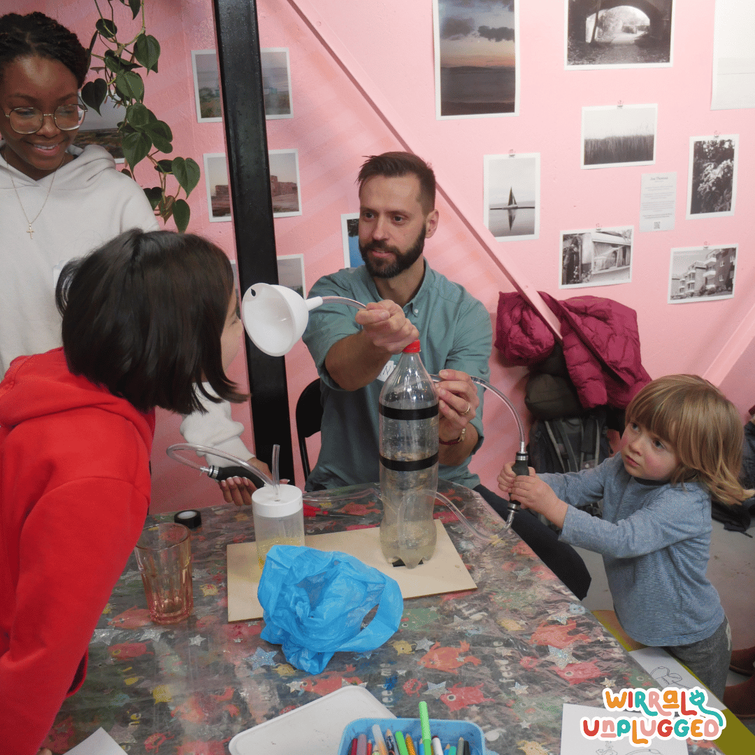 Chemist using a homemade industrial scrubber made from pop bottle and funnel . One child pumps the liquid and another one smells the funnel while a volunteer looks on smiling. Everything is low key and the group is inclusive.