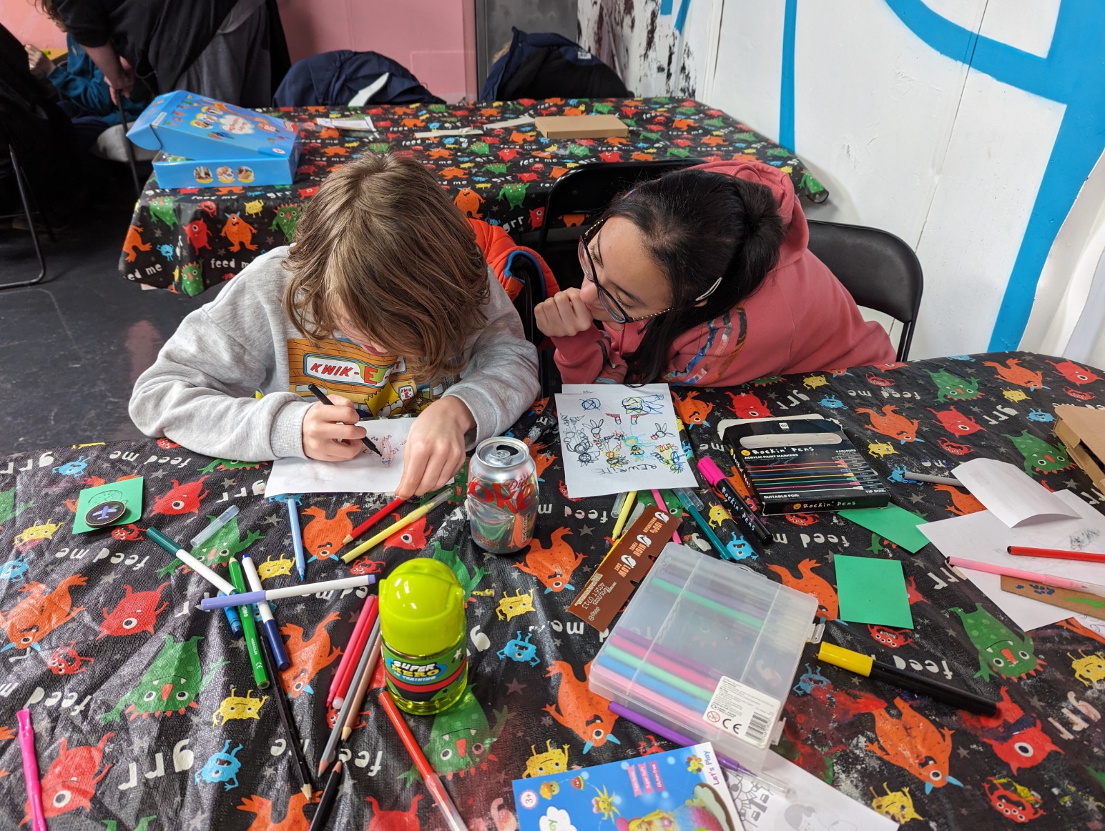 University student leans over young person drawing. The table cloth is brightly coloured with monsters and markers are spread out around the two.