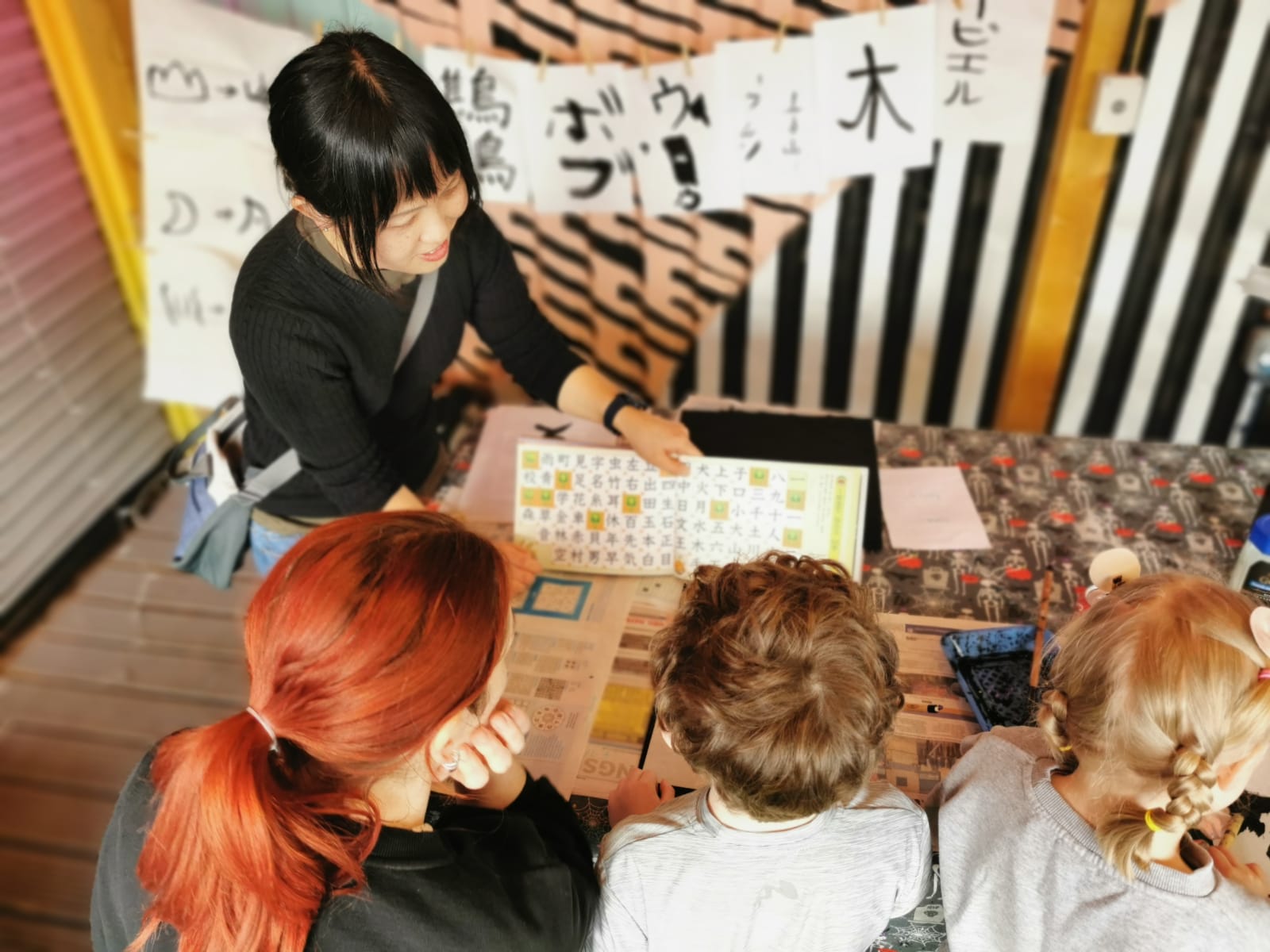Woman demonstrates Japanese calligraphy to a mum and her 2 young people, She's pointing at a book, and Ink drawings of previous people's calligraphy hang up behind her